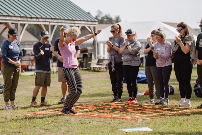 In the image, a woman is celebrating with her arms raised, while a group of people watches. They are standing on a grassy field with a geometric pattern marked on the ground. The people are dressed casually, and the scene appears to be outdoors on a sunny day.